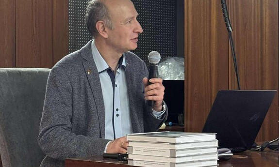 Mężczyzna w marynarce siedzi przy biurku, trzyma mikrofon i mówi do publiczności podczas prezentacji. Na biurku leży stos książek i otwarty laptop.