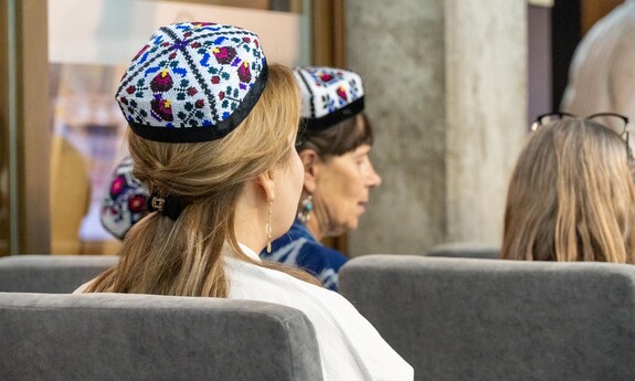 Kobiety siedzące wydarzeniu ubrane w tradycyjne haftowane czapeczki typu tubetejka z kolorowymi wzorami.