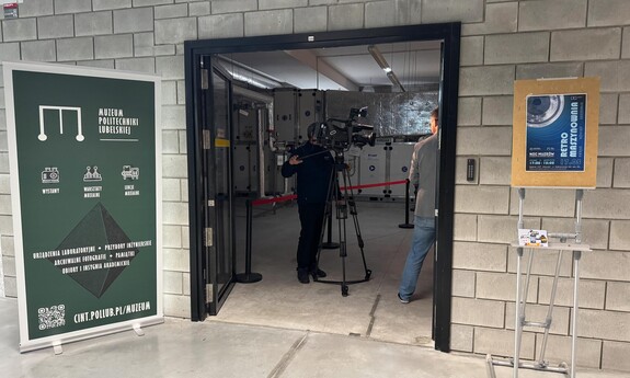 Wejście do Muzeum Politechniki Lubelskiej, przy którym ustawiono kamerę telewizyjną i trwa nagranie. Po lewej stronie widoczny zielony baner informacyjny muzeum, po prawej plakat wydarzenia „Retro Muzeum” z okazji Nocy Muzeów.