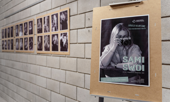 Galeria Centrum Informacji Naukowo-Technicznej Politechniki Lubelskiej, na pierwszym planie sztaluga z plakatem przedstawiającym zdjęcie kobiety siedzącej na kzreśle z aparatem przy twarzy, napisy: Centrum Informacji Naukowo-Technicznej Politechniki Lubelskiej zaprasza na wystawę z okazji Dnia Bibliotekarza i Bibliotek sami swoi portrety fotograficzne pracowników CINT,  zdjęcia: Marta Zbańska, Paulina Artymiuk-Broda, Katarzyna Krygier-Durakiewicz, w tle plansze ze zdjęciami 