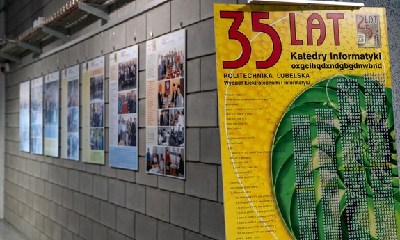 Widok wystawy z okazji 35-lecia Katedry Informatyki Politechniki Lubelskiej. Na pierwszym planie plakat z kolorową grafiką przedstawiający hasło '35 lat Katedry Informatyki' oraz kod binarny i fragmenty kodu w języku C. W tle na ścianie widoczne są plansze z tekstami i zdjęciami dokumentującymi historię i działalność katedry, rozmieszczone wzdłuż betonowej ściany korytarza.